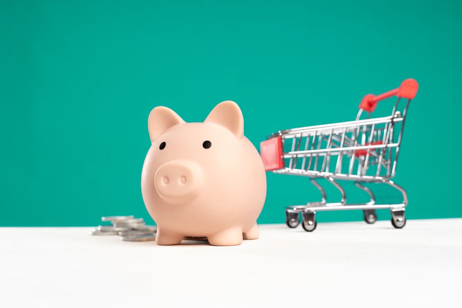 A person's hand carefully placing a coin into a white, house-shaped piggy bank against a bright, clean background, representing the concept of saving money on a home sale.